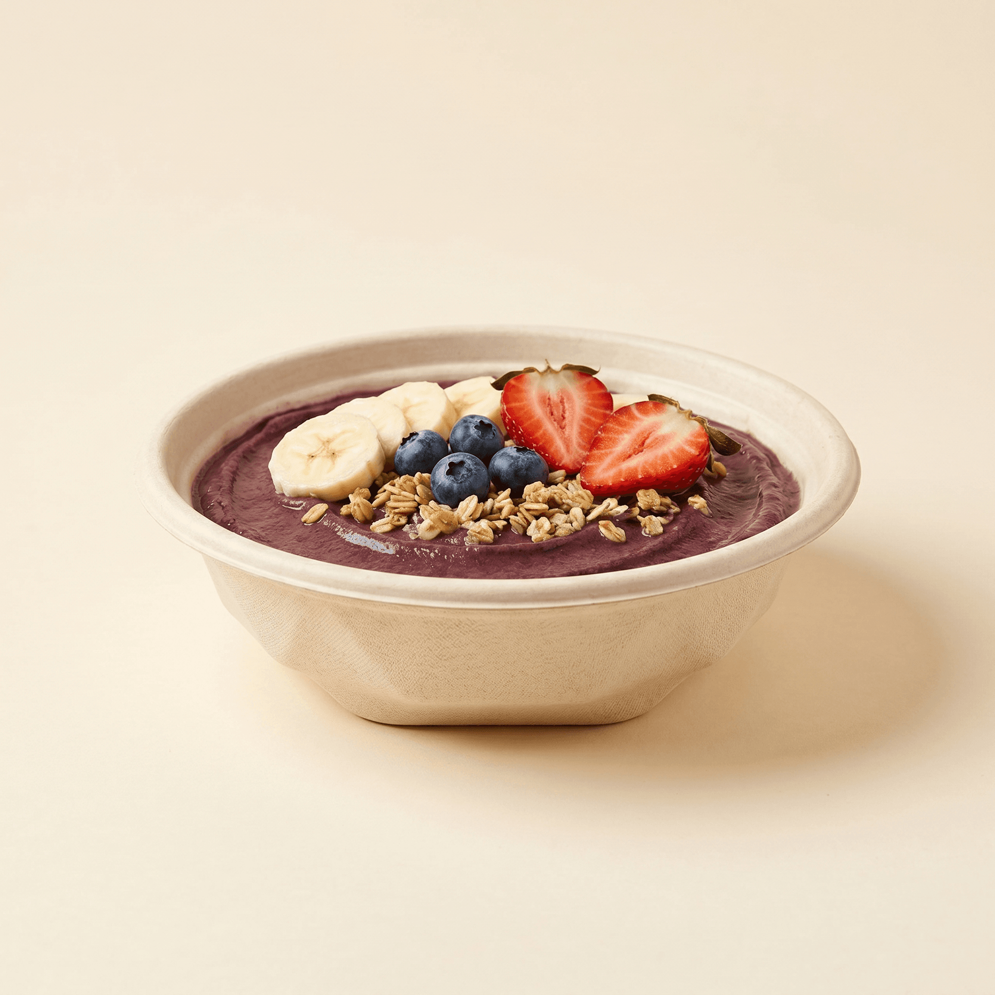 Bowl filled with a cold açaí bowl topped with banana, blueberries, strawberries and granola on a cream studio background, showing it holds cold food