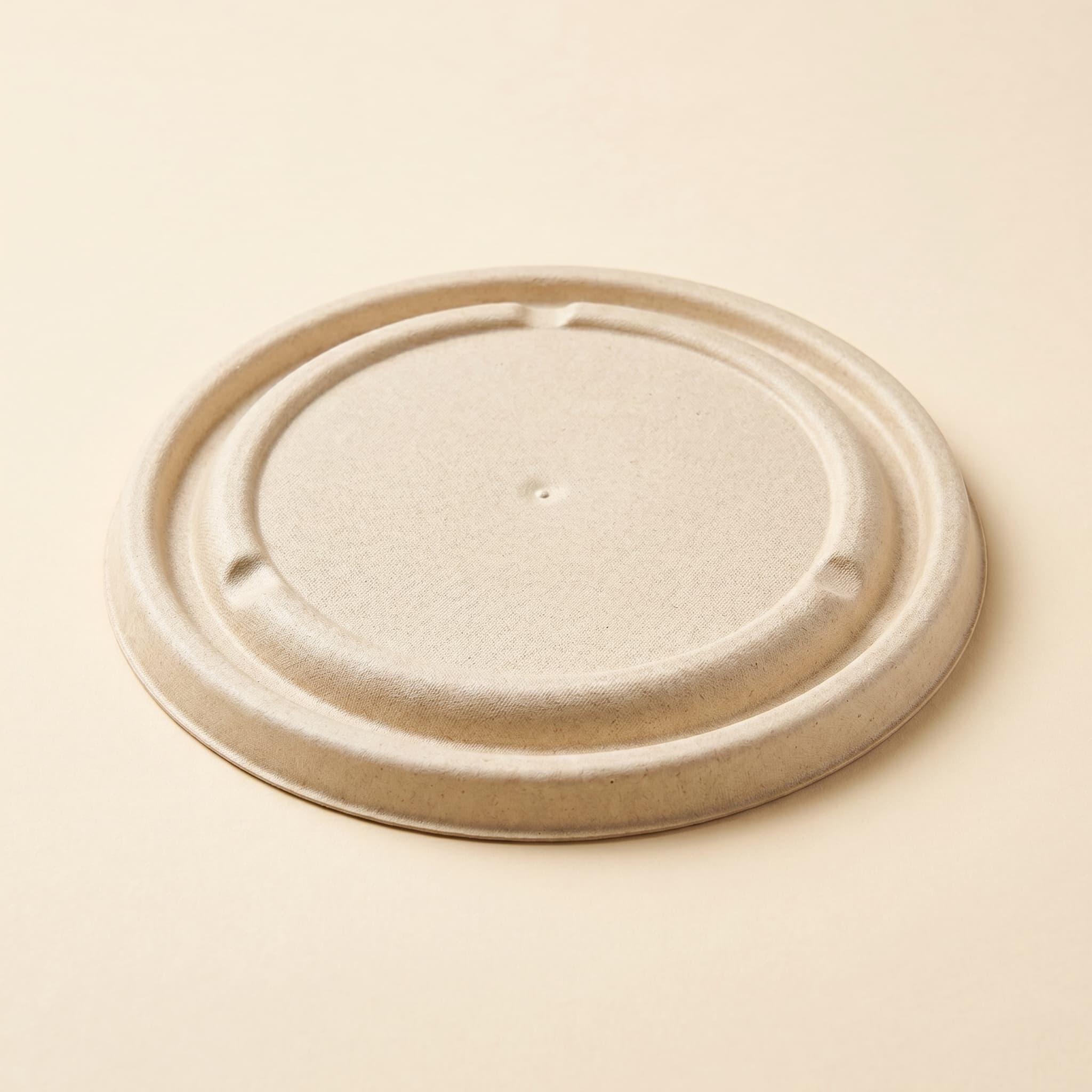 Molded sugarcane bagasse fiber bowl lid at a three-quarter angle on a cream studio background, showing the low domed top, concentric grooves and rolled snap-over rim